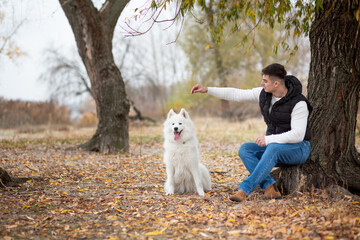 A guy trains his pet Samoyed while sitting in a park by the river