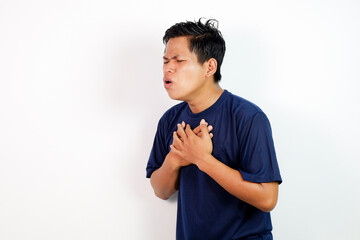 A young Asian man in a blue shirt clutches his chest with a pained expression, conveying distress or chest discomfort in a simple studio setting.