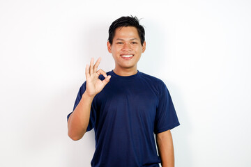 A cheerful young Asian man in a blue shirt stands against a clean white background, smiling and giving an OK gesture.