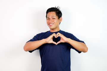 A cheerful young Asian man in a navy blue shirt forms a heart shape with his hands against a clean white background, conveying love, friendship, positivity, and healthy lifestyle.