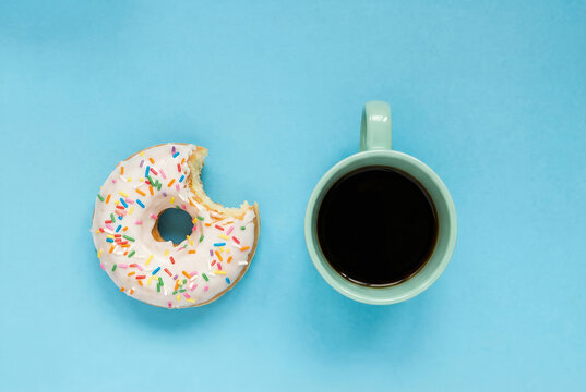 Minimalist flat lay of a bitten white glazed donut with colorful sprinkles and a cup of black coffee. Top view breakfast concept on a pastel blue background - Powered by Adobe