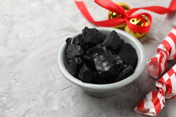 Pieces of black coal in bowl, candies and Christmas jingle bells on gray textured table, closeup. Space for text