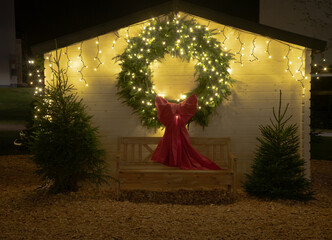A large glowing Christmas wreath with a red bow decorates a small wooden hut, framed by fairy lights and festive evergreen trees.