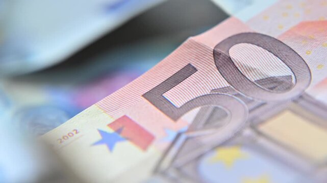 Euro banknotes being inspected with a magnifying glass. The magnification highlights intricate security features, micro-printing, and detailed textures, creating an analytical financial scene