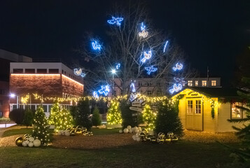 A festive Christmas park scene with glowing trees, illuminated angel decorations, ornaments, and a warmly lit holiday cabin at night.