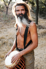 Smiling bearded hippie with long hair holding djembe in golden forest light