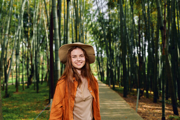 Obraz premium Woman in a straw hat with a warm smile in a bamboo forest on a wooden path, portrait of a happy traveler wearing orange jacket, nature walkway with sunlight through tall green stalks.
