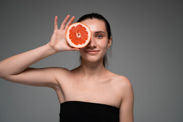 Positive young woman holding a grapefruit slice over her eye, highlighting citrus freshness, natural skincare