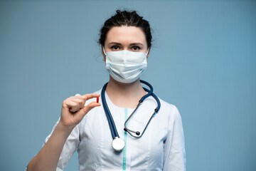 Female doctor in medical mask showing red pill toward camera while wearing white uniform
