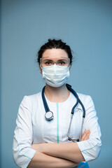 Medical doctor in mask and protective eyewear posing with crossed arms on blue hospital background