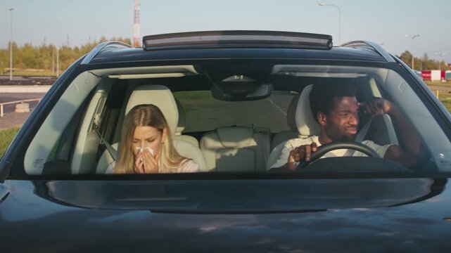 Couple inside car sneezing and coughing into tissues during drive. Woman holding napkin covering face. Man behind wheel bending forward sick. Chilly morning light outside road background.