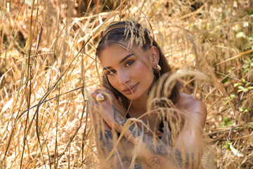 Portrait of a young, beautiful, Latina woman with tattoos, wearing a white crochet dress, intimate and tender, connected to nature and ecology, sitting on the ground surrounded by dry grasses.