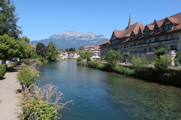 Fototapeta premium Riverfront view of buildings and mountains under a clear blue sky, summer