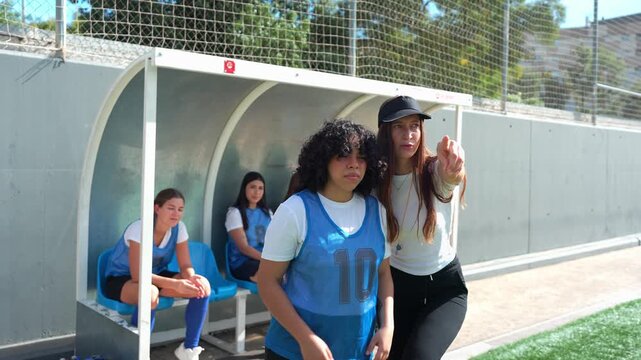 Young female coach explaining tactics to a player before she enters the field. Teammates watch from the bench during a competitive game