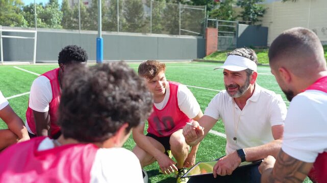 Professional male coach explaining game tactics and strategy to his multiethnic team. Players are attentively listening on the field