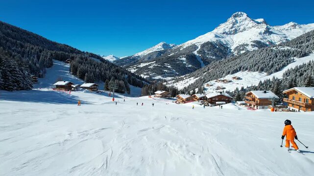 Child skier on snowy mountain slope with chalets and pine trees on a sunny day