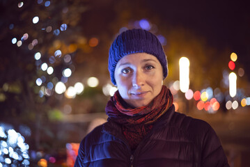 A close-up portrait of a woman (30s) wearing a hat and scarf, smiling directly at the camera with a brilliant, colored bokeh background. Focuses on the cheerful mood and the warm illumination
