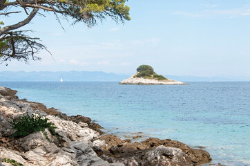 Rocky coastline with clear turquoise water, a small tree-covered island, distant mountains, and a calm summer seascape under a bright sky. Nature landscape, travel and vacation destination by the sea.