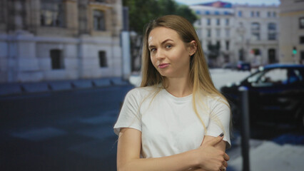 Fototapeta premium Woman standing on city street outdoors with arms crossed wearing casual white shirt near urban buildings on a clear day, showcasing a thoughtful expression and long hair.