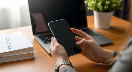 Person using smartphone and laptop at wooden desk with notebook and plant.