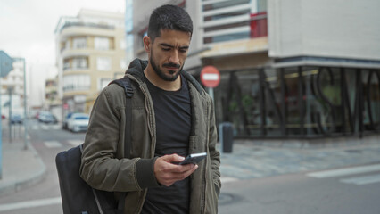 Young man using smartphone on urban street, wearing a jacket and carrying backpack, surrounded by...