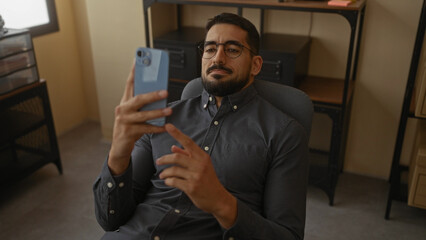 Young man with beard relaxing in an office chair while using smartphone indoors, surrounded by shelves and storage boxes, showcasing a modern workplace environment.