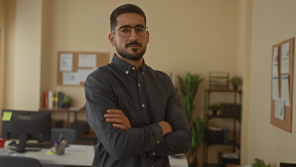 Young man with beard and glasses adjusting shirt and crossing arms in modern office setting,...