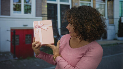 Woman holds pink gift box tied with ribbon in front of her chest on street and smiles with delight; joy.