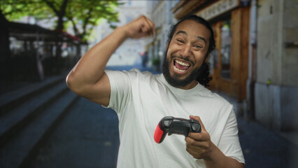 Man holding black and red game controller with both hands and raising his right fist while looking at camera on a city street lined with shops and steps; excitement.