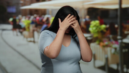 Woman covering face with hands in outdoor restaurant terrace highlighting emotion of fear amidst lively background.