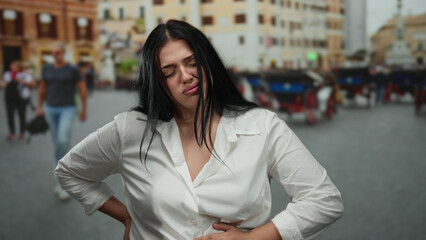 Fototapeta premium Woman in white shirt with stomach pain stands on a busy city street surrounded by blurred pedestrians and buildings under daylight.