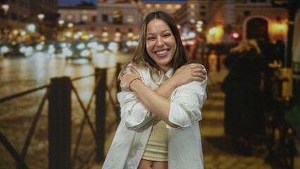 Woman smiling and hugging herself on a wet cobblestone city street lined with lights at night; self care wellness joy.