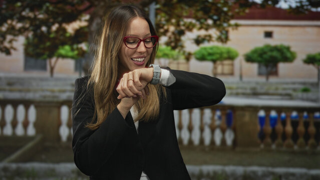 Woman checking smartwatch on wrist by stone balustrade in front of building, wearing red glasses and black blazer and smiling while looking at time; time management optimism. - Powered by Adobe