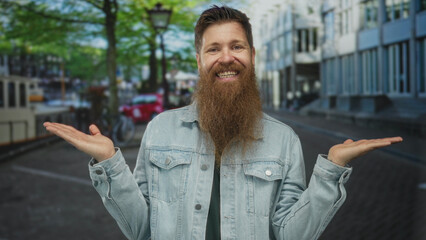 Man in denim jacket gently gestures open palms and smiles on sunlit brick paved city street;...