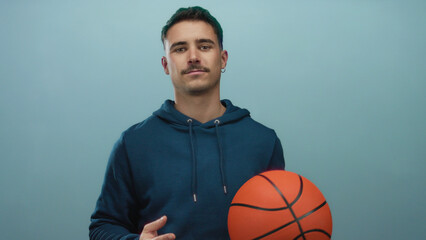 Young hispanic man holding basketball against isolated blue background with wall, embodying sporty casual athletic look, symbolizing urban youthful energy and focus