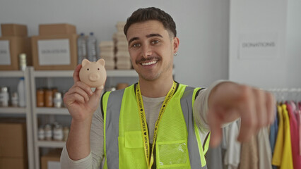 Young man with reflective vest holding piggy bank in a charity donations center surrounded by boxes and clothes, symbolizing voluntary support and aid.