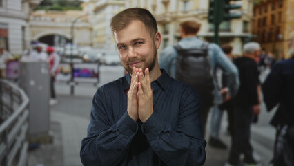 Naklejka premium Young caucasian man in navy shirt clasping hands and smiling on a busy city street near pedestrians and historic buildings; playful.