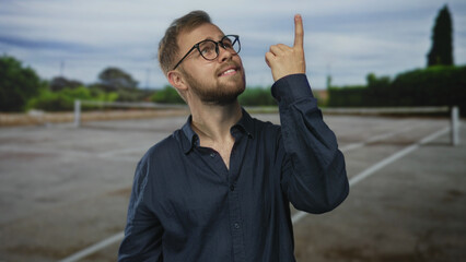 Naklejka premium Man with glasses pointing index finger up on a street parking area while looking upward; hope curiosity.