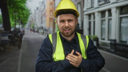Man builder clutching hands to chest on city street wearing yellow hardhat and neon safety vest; medical emergency distress.