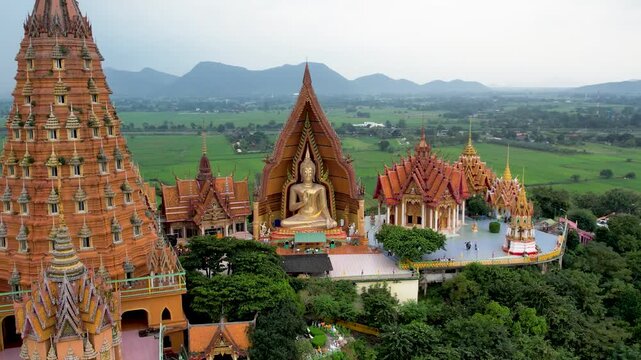 An aerial drone view of Wat Tham Suea temple in Kanchanaburi, Thailand, surrounded by vast green rice fields and distant mountains.