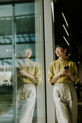 Woman standing by glass window smiling while holding smartphone in modern cafe setting