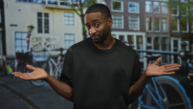 Man in black t shirt shrugging with hands raised on a city street near bicycles and canal buildings, bare forearms visible; uncertainty.