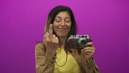 Smiling woman holding retro camera and showing middle finger with direct gaze in purple studio; defiance.