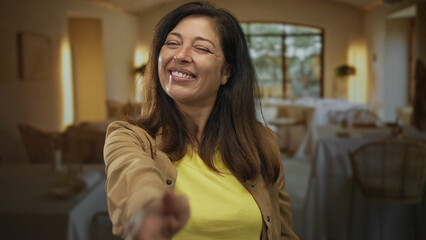 Woman in yellow shirt seated outdoors smiles and extends hand forward in restaurant building;...