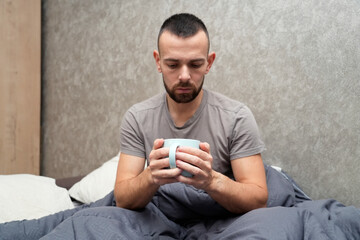 A man sits in bed holding a mug, looking tired and unwell. He is resting under a blanket, taking time off work to recover from an illness and regain his health