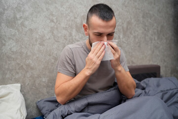 A man sits up in bed looking tired and unwell. He is using tissues to manage his symptoms. The room is simple and indicates he is taking time off for sickness and recovery