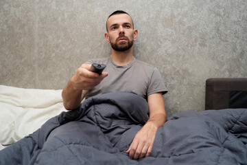 A man sits up in bed, wrapped in a blanket, holding a TV remote. He looks tired and is taking time off work to rest and recover from illness at home