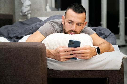 A man lies on a bed with a pillow, looking at his phone. He appears to be resting while recovering from illness. The setting is indoor. The atmosphere suggests fatigue and a need for rest