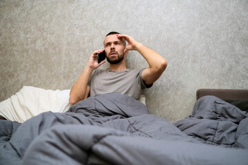 A man lies in bed covered with a blanket, appearing fatigued. He talks on the phone, seeking help or sharing updates while resting in his bedroom at home during the day