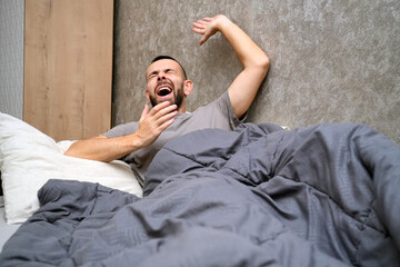A man lies in bed under a blanket, yawning widely. He appears to be experiencing fatigue and is taking time to recover from an illness at home in a simple room